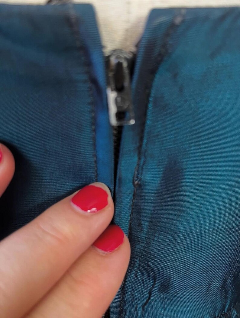 May include: Close-up of teal fabric with a silver zipper. The fabric has a slight sheen, possibly from a jacket or coat. The zipper is partially closed, showing the fabric's texture and colour.