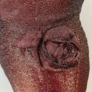 May include: A close-up of a burgundy sequined fabric with a large rose-shaped embellishment. The fabric has a subtle shimmer and the rose is made of the same material.