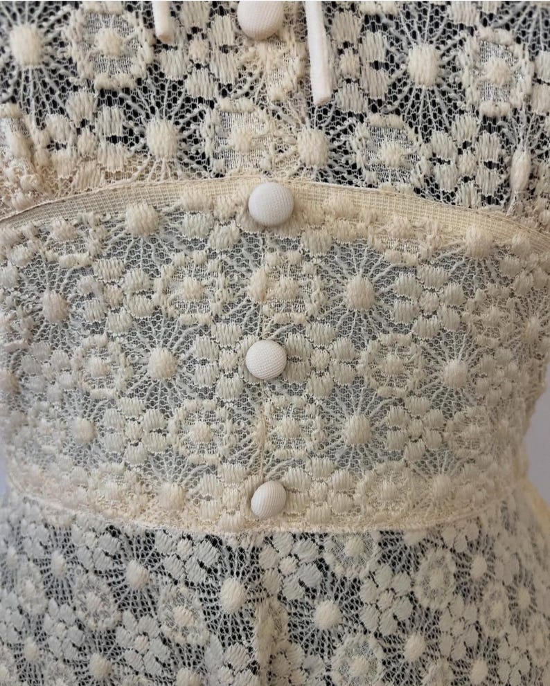 May include: Close-up of a cream-colored lace fabric with a floral pattern. The fabric has a series of white buttons down the center.