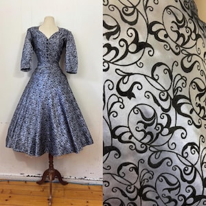 May include: A vintage blue dress with a black floral pattern. The dress features a V-neckline, three-quarter sleeves, and a flared skirt. It is displayed on a wooden dress form, showcasing its classic silhouette and design.