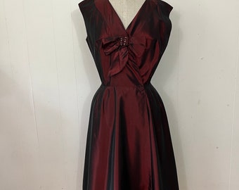 May include: A sleeveless, knee-length dress in a burgundy and black color scheme. The dress features a V-neckline with a bow detail at the bust and a flared skirt. The dress is displayed on a mannequin against a white background.