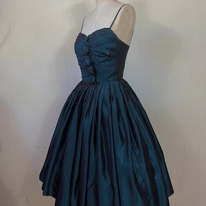 May include: A teal vintage dress on a mannequin. The dress has a fitted bodice with spaghetti straps and a gathered, ruched design. The full skirt flares out, creating a classic silhouette. The dress is made of a shiny fabric.