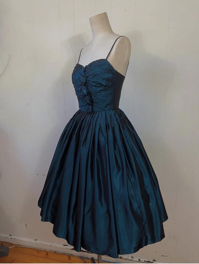 May include: A teal vintage dress on a mannequin. The dress has a fitted bodice with spaghetti straps and a gathered, ruched design. The full skirt flares out, creating a classic silhouette. The dress is made of a shiny fabric.