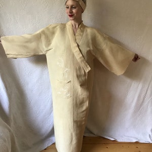 Puede incluir: Una larga bata de lino de color crema con un diseño bordado en blanco. La bata tiene una silueta amplia y fluida y un cinturón para atar a la cintura.