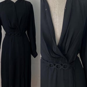Puede incluir: Un vestido negro de manga larga con un corpiño cruzado y una falda a la rodilla. El vestido presenta un cinturón decorativo con tres acentos circulares en la cintura. El vestido se exhibe en un maniquí.