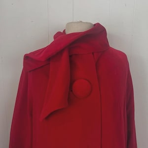 May include: A vibrant red coat with a unique scarf-like collar and large, matching buttons. The coat is displayed on a mannequin, showcasing its design and style. The buttons are circular and add a classic touch to the overall look.