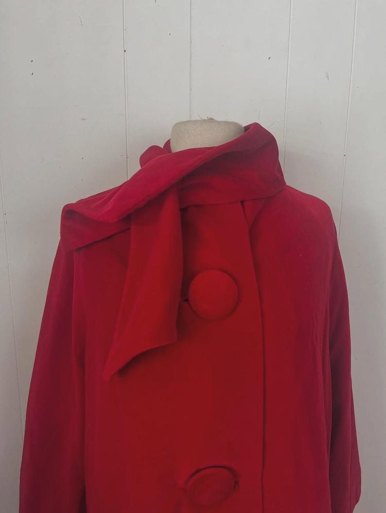 May include: A vibrant red coat with a unique scarf-like collar and large, matching buttons. The coat is displayed on a mannequin, showcasing its design and style. The buttons are circular and add a classic touch to the overall look.
