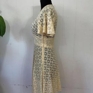 May include: A cream-colored lace dress with a V-neckline and short, flared sleeves. The dress has a fitted bodice and a flowing skirt.