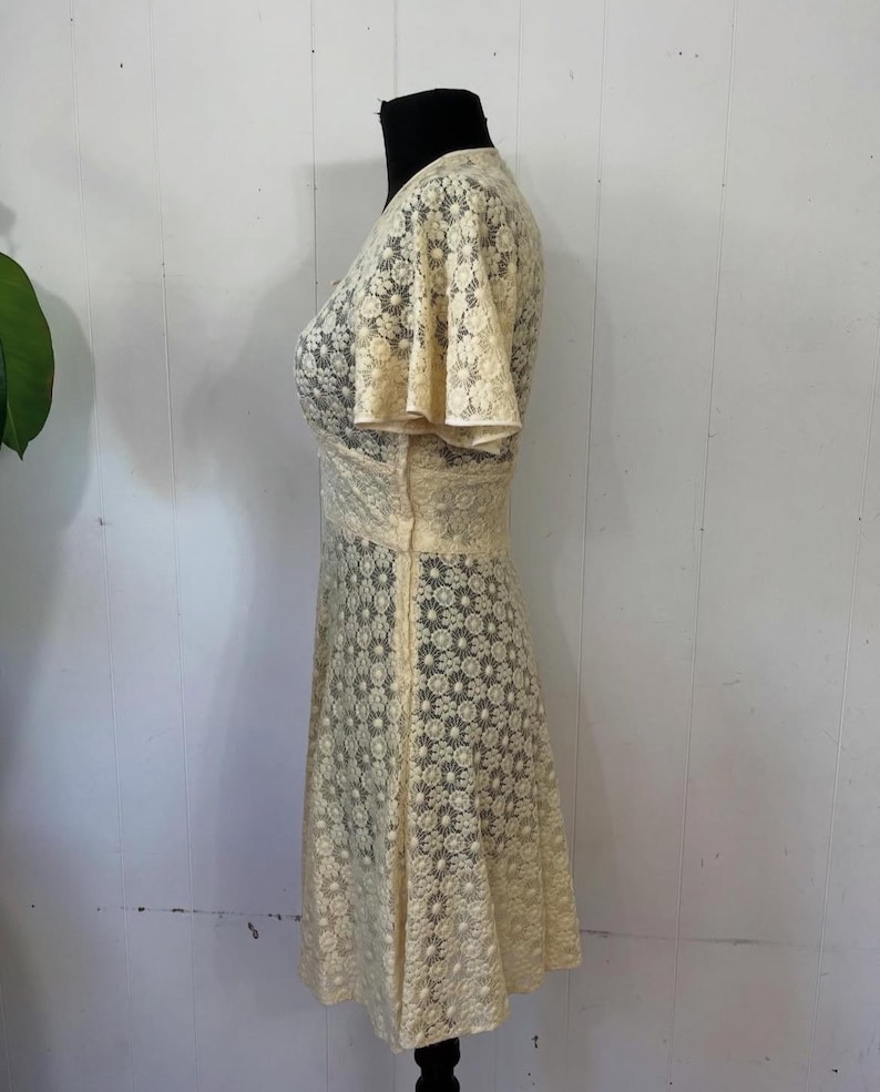 May include: A cream-colored lace dress with a V-neckline and short, flared sleeves. The dress has a fitted bodice and a flowing skirt.
