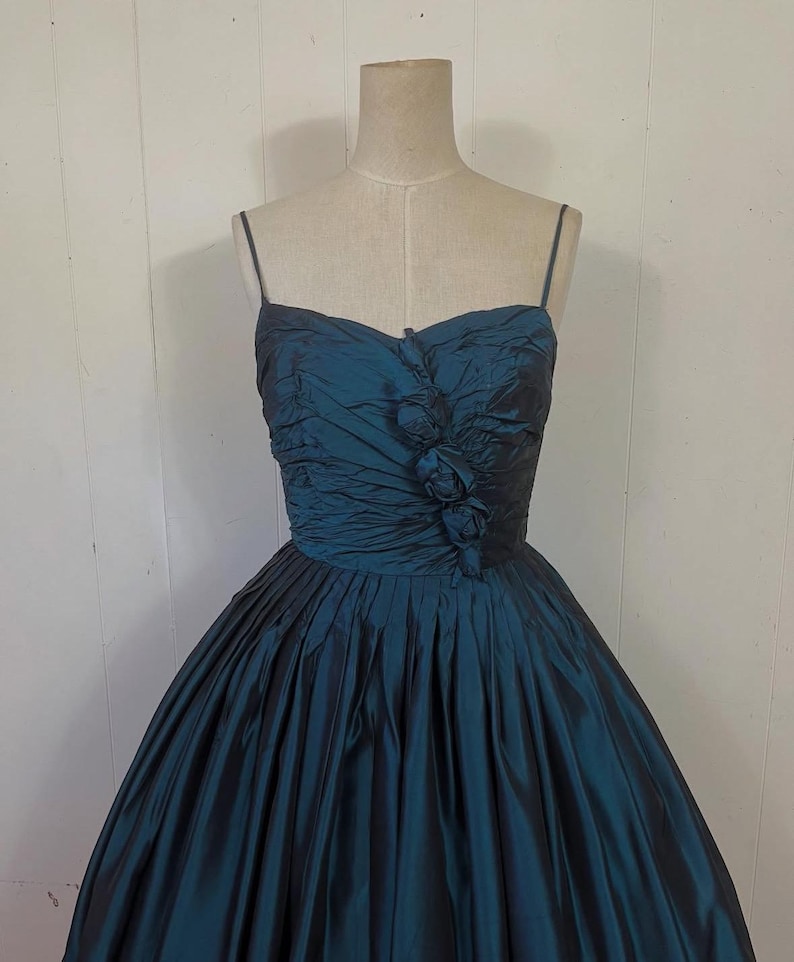 May include: A teal vintage dress with a sweetheart neckline and spaghetti straps. The bodice is ruched with floral detailing, and the full skirt is pleated. The dress is displayed on a mannequin.