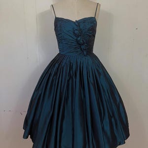 May include: A teal vintage dress with a sweetheart neckline and spaghetti straps. The bodice is ruched and has floral detailing. The full skirt flares from the waist, creating a classic silhouette. The dress is displayed on a mannequin.