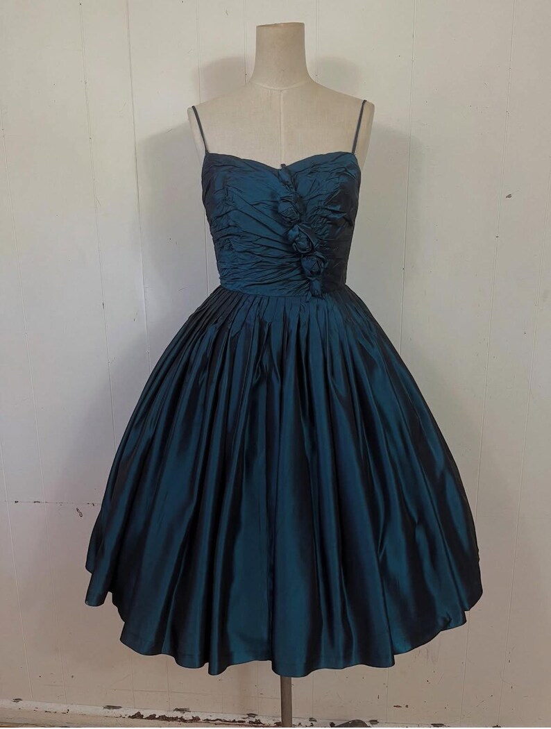 May include: A teal vintage dress with a sweetheart neckline and spaghetti straps. The bodice is ruched and has floral detailing. The full skirt flares from the waist, creating a classic silhouette. The dress is displayed on a mannequin.