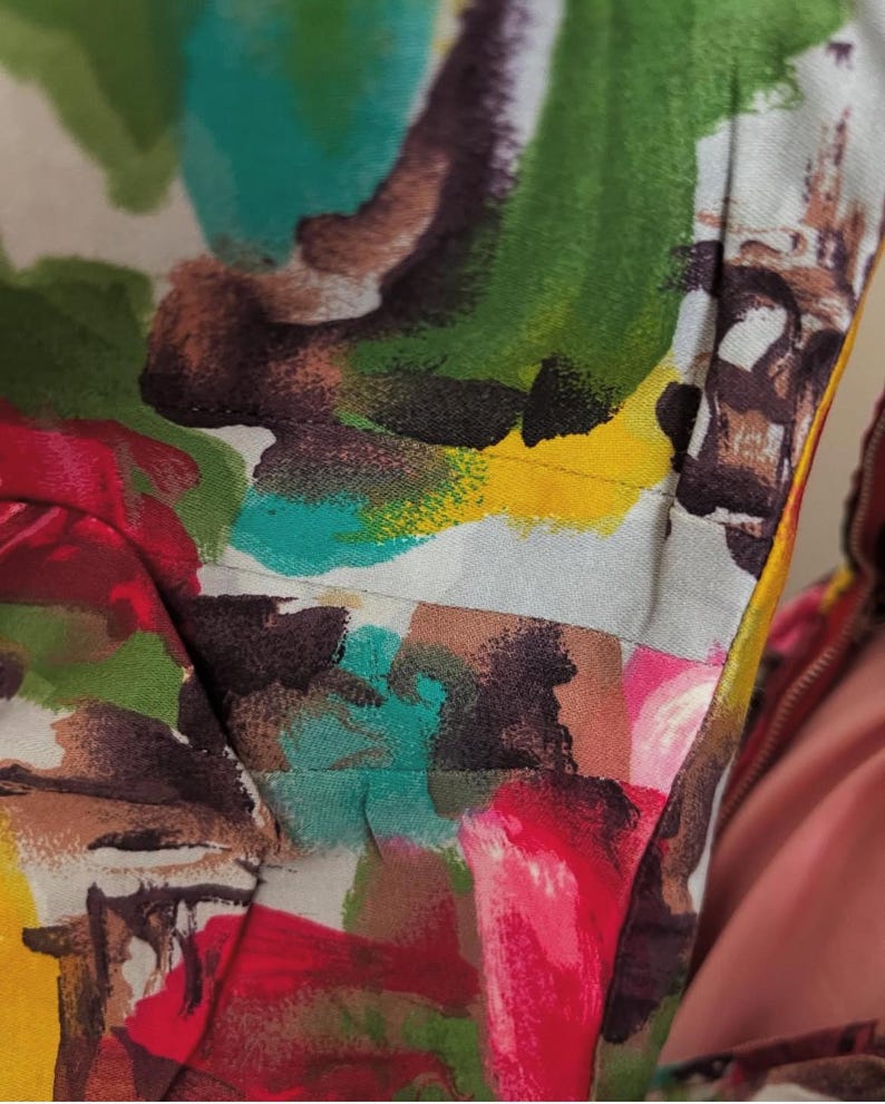 May include: Close-up of a colorful fabric with abstract brushstrokes in shades of red, pink, green, blue, yellow, and brown. The fabric has a white background and a subtle texture.