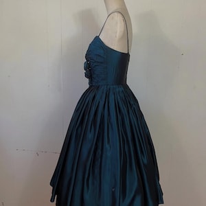 May include: A teal vintage dress with a fitted bodice and a full skirt. The dress has spaghetti straps and floral embellishments on the bodice. The fabric has a sheen, possibly silk or satin. The dress is displayed on a mannequin. The dress is a classic vintage style.