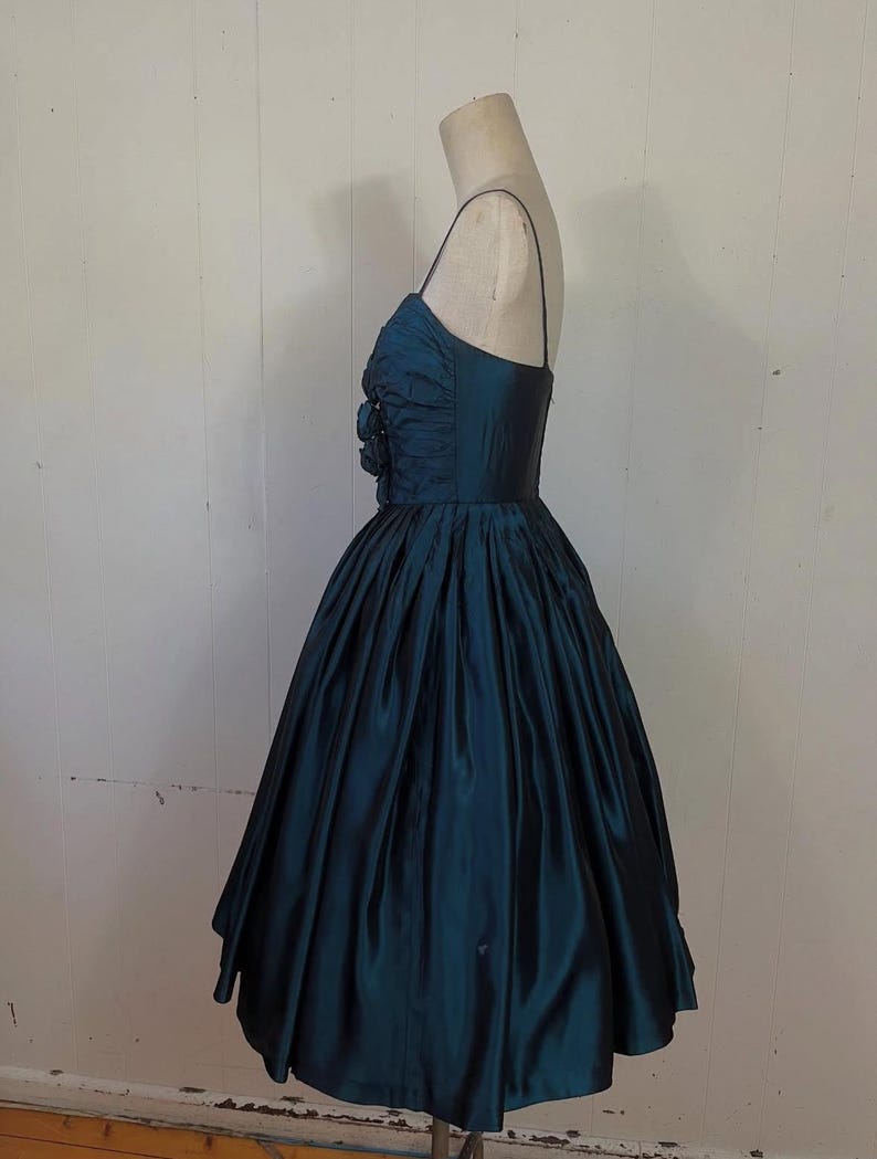 May include: A teal vintage dress with a fitted bodice and a full skirt. The dress has spaghetti straps and floral embellishments on the bodice. The fabric has a sheen, possibly silk or satin. The dress is displayed on a mannequin. The dress is a classic vintage style.