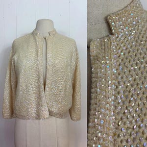 May include: Cream-coloured, open-front cardigan with a high collar. The vintage sweater is covered in iridescent sequins, creating a shimmering effect. The long-sleeved garment is displayed on a mannequin.