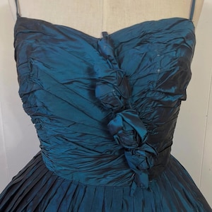 May include: A teal vintage dress with a pleated skirt and a ruched bodice. The dress has delicate spaghetti straps and fabric roses at the bust. The fabric has a subtle sheen, adding to its elegant appearance.