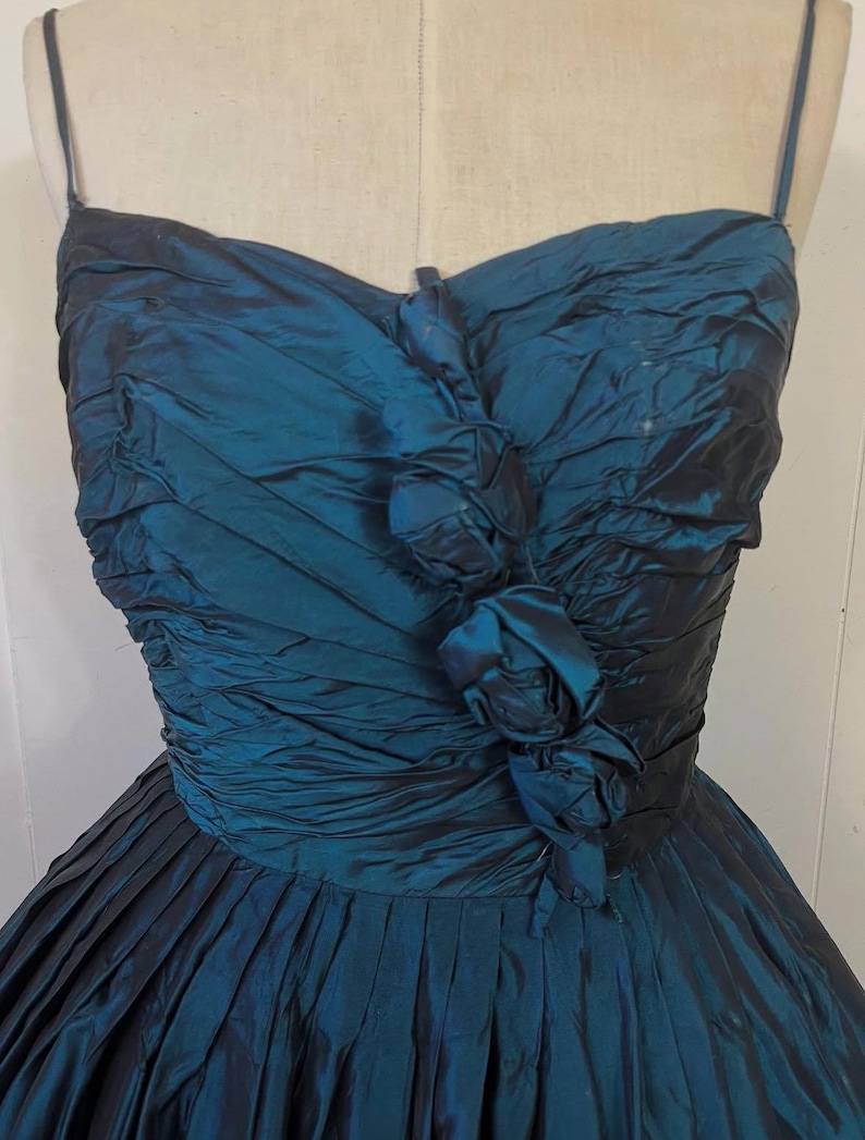 May include: A teal vintage dress with a pleated skirt and a ruched bodice. The dress has delicate spaghetti straps and fabric roses at the bust. The fabric has a subtle sheen, adding to its elegant appearance.