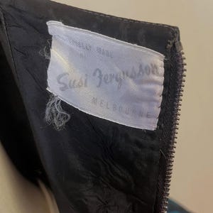 May include: Close-up of a black garment with a zipper and a white label. The label reads "Susi Bergsson MELBOURNE" in cursive script. The garment's fabric appears to be lined with a contrasting material.