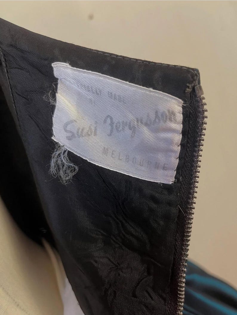 May include: Close-up of a black garment with a zipper and a white label. The label reads "Susi Bergsson MELBOURNE" in cursive script. The garment's fabric appears to be lined with a contrasting material.