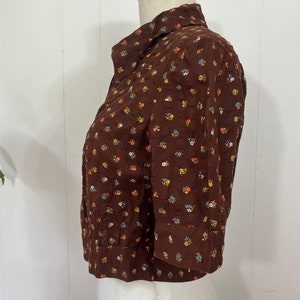 May include: A brown button-up shirt with a small floral print in shades of blue, orange, and yellow. The shirt has a collared neckline and three-quarter sleeves.