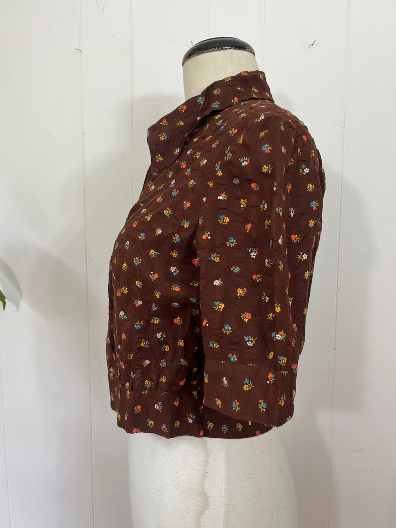 May include: A brown button-up shirt with a small floral print in shades of blue, orange, and yellow. The shirt has a collared neckline and three-quarter sleeves.