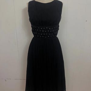 May include: A sleeveless black dress displayed on a mannequin. The dress features a fitted bodice with a beaded waistline and a pleated skirt that falls to the mid-calf. The dress is made of a lightweight fabric.