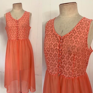 May include: A vintage coral-colored dress displayed on a mannequin. The sleeveless dress features a V-neckline and a bodice with a floral lace overlay. The skirt is a flowing, semi-sheer fabric. The dress is buttoned down the front.
