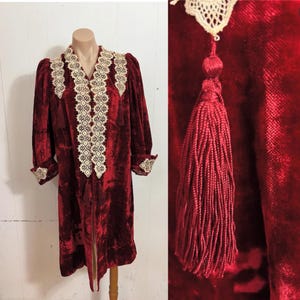 May include: A burgundy velvet robe with cream-colored lace trim and a matching tassel. The robe features long sleeves, a V-neck, and decorative lace down the front. The robe is displayed on a mannequin.