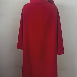 May include: A long, vibrant red coat with a high collar and long sleeves. The coat is made of a smooth fabric and appears to be a vintage or costume piece. The coat is displayed on a wooden stand against a white wall.