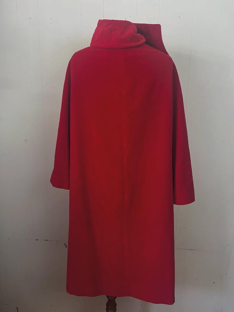May include: A long, vibrant red coat with a high collar and long sleeves. The coat is made of a smooth fabric and appears to be a vintage or costume piece. The coat is displayed on a wooden stand against a white wall.