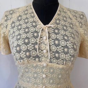 May include: A cream-colored lace dress with a V-neckline and short sleeves. The dress has a tie at the neckline and buttons down the front.