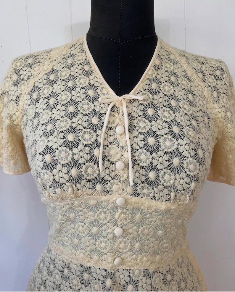 May include: A cream-colored lace dress with a V-neckline and short sleeves. The dress has a tie at the neckline and buttons down the front.