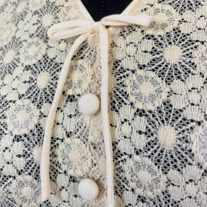 May include: Close-up of a cream-colored lace top with a bow tie and buttons. The lace has a floral pattern.
