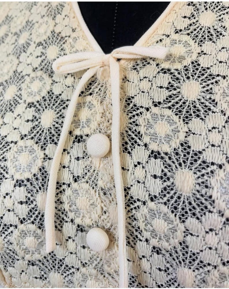 May include: Close-up of a cream-colored lace top with a bow tie and buttons. The lace has a floral pattern.