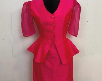 May include: A vibrant fuchsia two-piece suit. The jacket features a V-neck, button closure, and a peplum waist. The sleeves are short with sheer, puffed fabric. The matching pencil skirt completes the ensemble. The suit is displayed on a black mannequin.