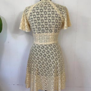 May include: A cream-colored lace dress with a floral pattern. The dress has a V-neckline and short sleeves. The skirt is a-line and falls to just above the knees.