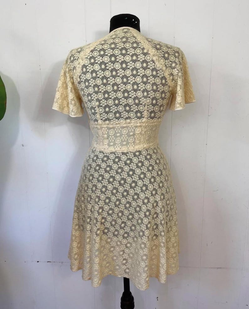 May include: A cream-colored lace dress with a floral pattern. The dress has a V-neckline and short sleeves. The skirt is a-line and falls to just above the knees.