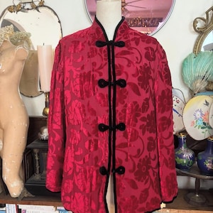 May include: A red floral patterned jacket with black trim and frog closures. The jacket is displayed on a mannequin. The jacket has a mandarin collar and long sleeves. The floral pattern is a darker shade of red.