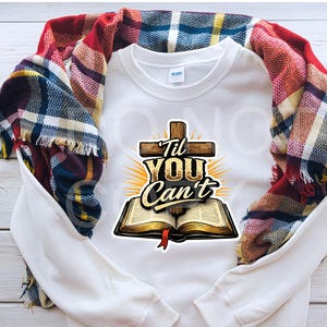Puede incluir: Sudadera blanca con un gráfico que presenta una cruz de madera, el texto "Til You Can't" y un libro abierto con una cinta roja. Una bufanda a cuadros en tonos rojos, azules y amarillos está sobre la sudadera.