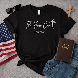 Puede incluir: Camiseta negra con el texto "Til You Can" y una cruz, con el nombre "Kid Rock" debajo. La camiseta se combina con gafas de sol, una bandera americana, una cruz de madera, una Biblia y vaqueros azules.