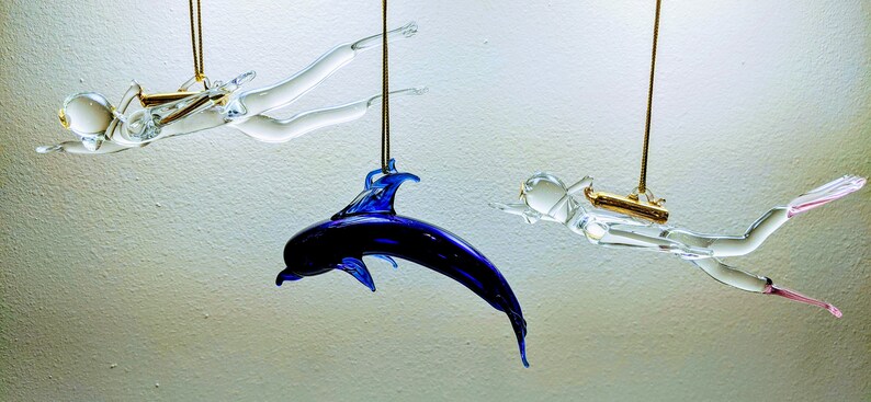May include: Three glass ornaments hanging from the ceiling. Two are clear glass figures of divers, one with a pink fin and the other with a gold tank. The third ornament is a blue glass dolphin.