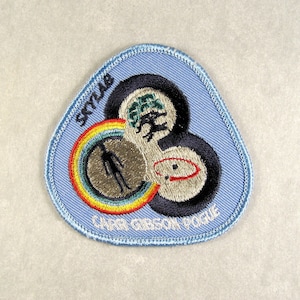 Puede incluir: Parche bordado de la misión Skylab con un fondo azul claro y una forma triangular única. El parche presenta tres diseños circulares interconectados con un arcoíris, una figura de astronauta y el texto "SKYLAB" y "CARR GIBSON POGUE."