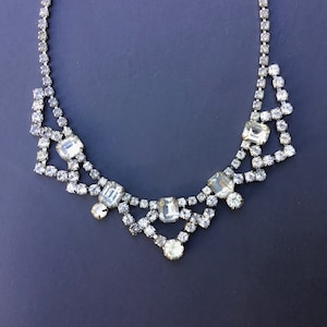 Puede incluir: Un collar plateado con brillantes pedrería transparente y brillante. El collar presenta una delicada cadena y una serie de formas geométricas adornadas con piedras claras y facetadas de varios tamaños. El diseño incluye piedras rectangulares y redondas.