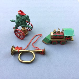 May include: Three Christmas ornaments: a green horse-drawn carriage with red wheels, a red and green wooden train, and a brass bugle with a red string. The ornaments are festive and decorative.