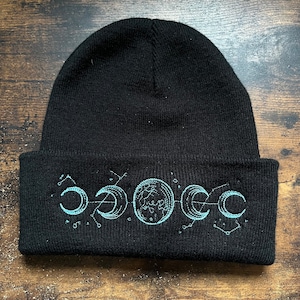 Könnte beinhalten: Schwarze Mütze mit Umschlag. Die Mütze hat ein hellblaues, gesticktes Design der Mondphasen. Das Design umfasst Halbmonde, Kreise und Verbindungslinien.