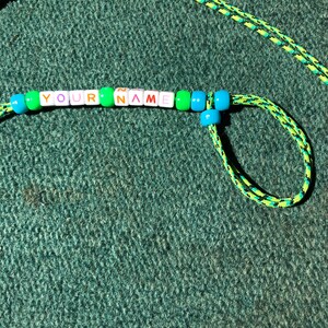 May include: A green and black lanyard with a beaded name tag that reads "YOUR NAME" in colorful letters. The beads are green, blue, pink, and orange.