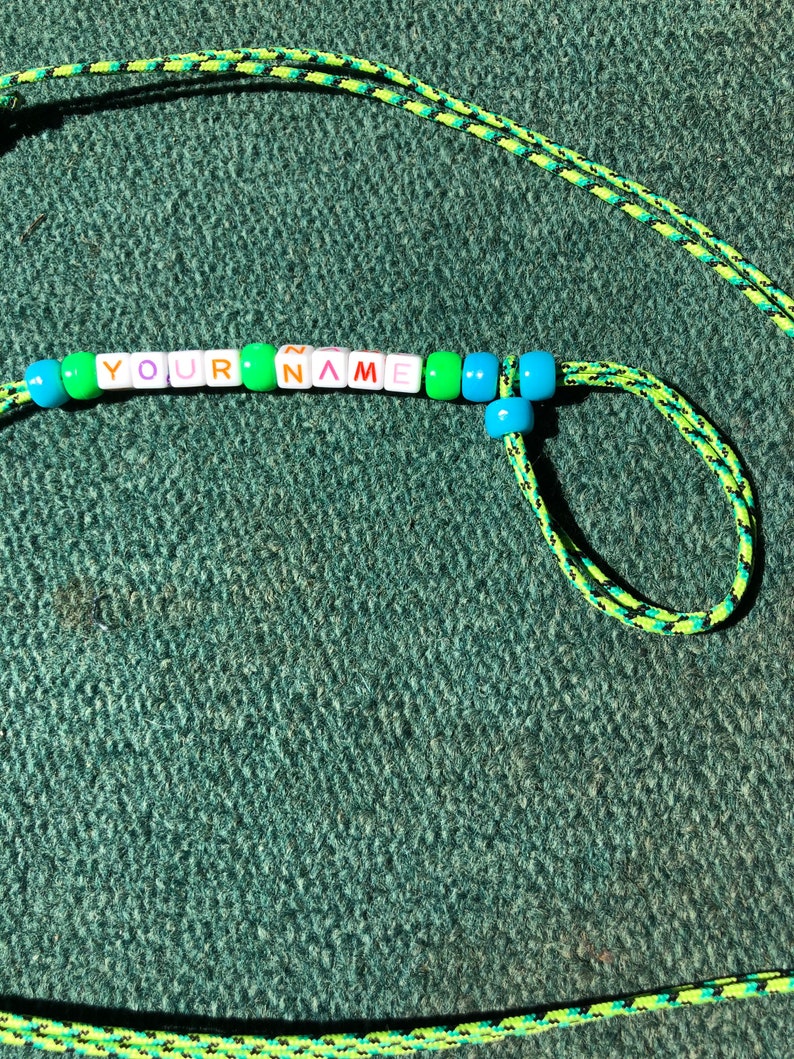 May include: A green and black lanyard with a beaded name tag that reads "YOUR NAME" in colorful letters. The beads are green, blue, pink, and orange.