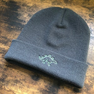 A dark grey beanie with the outline of a dinosaur embroidered onto it in green.