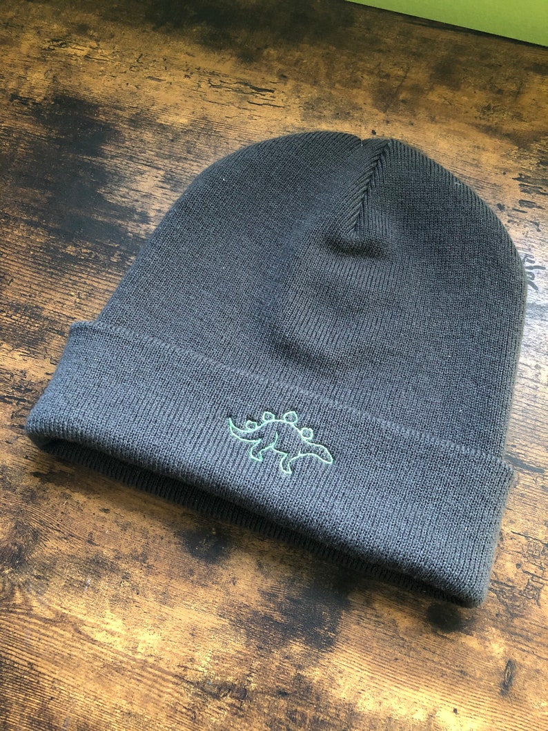 A dark grey beanie with the outline of a dinosaur embroidered onto it in green.